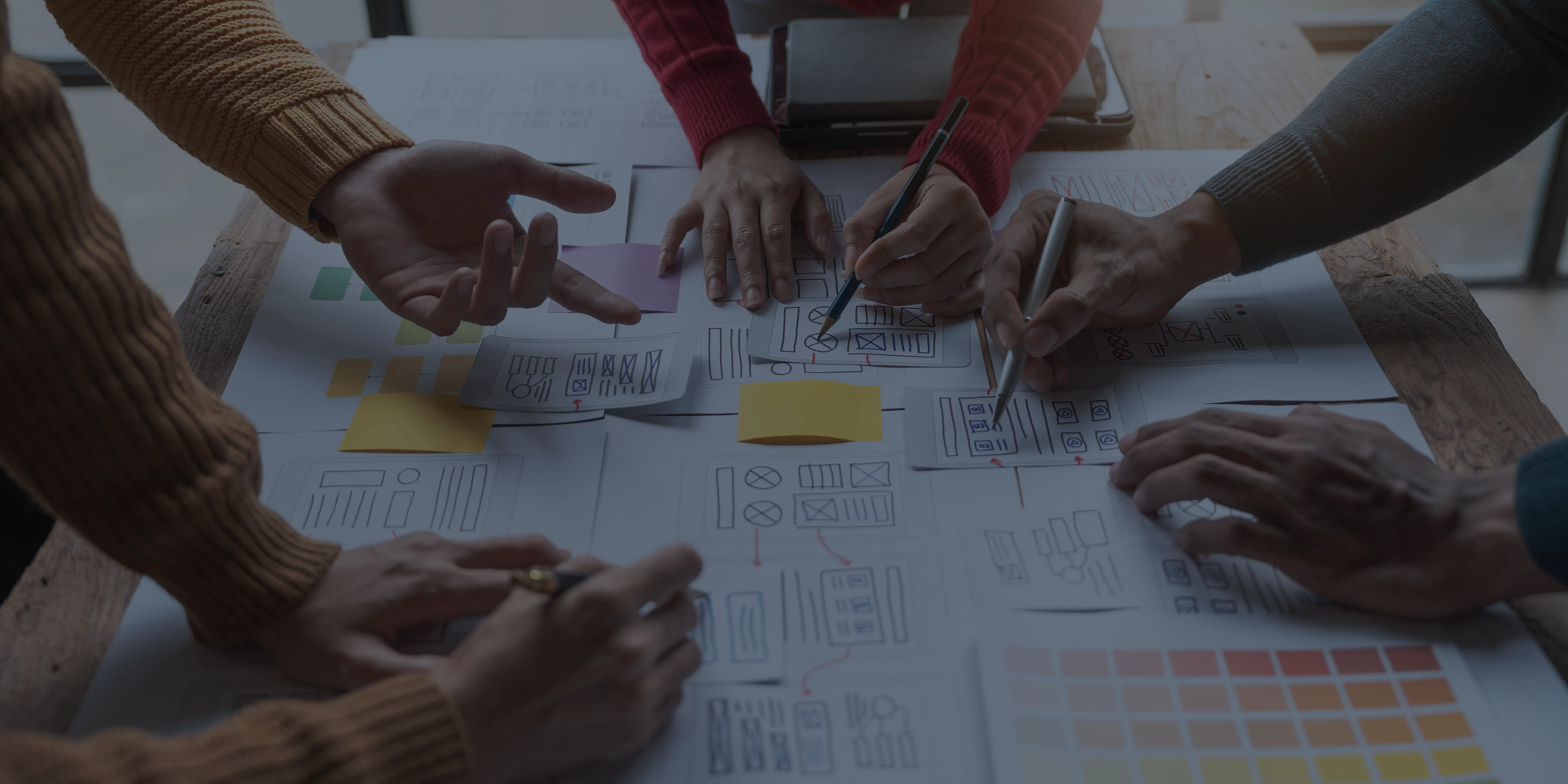 Multiple hands collaborating over a table filled with website wireframe sketches and sticky notes.