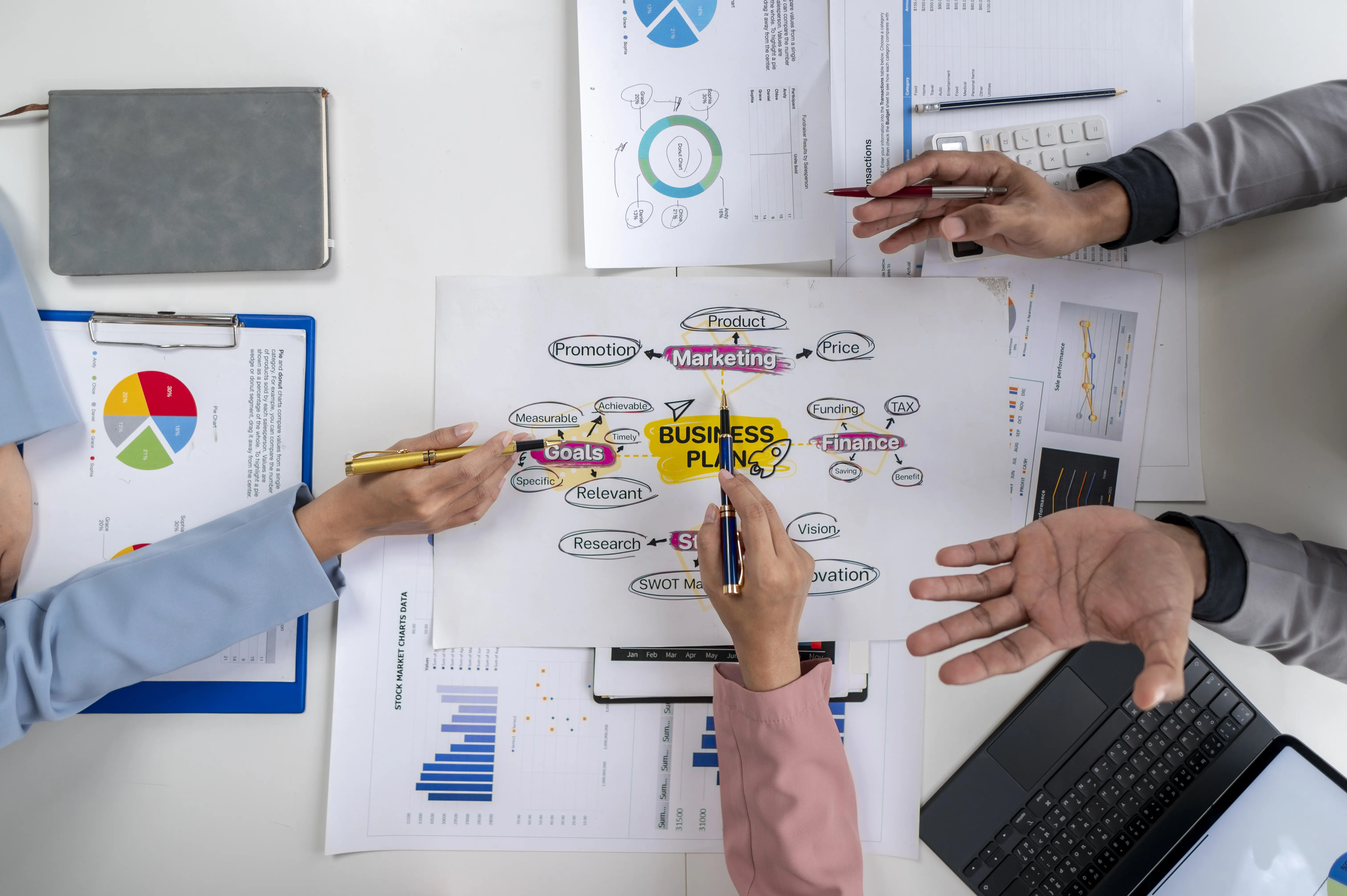 Overhead view of three people collaborating on a business plan diagram with charts, a laptop, and a notebook on a white table.