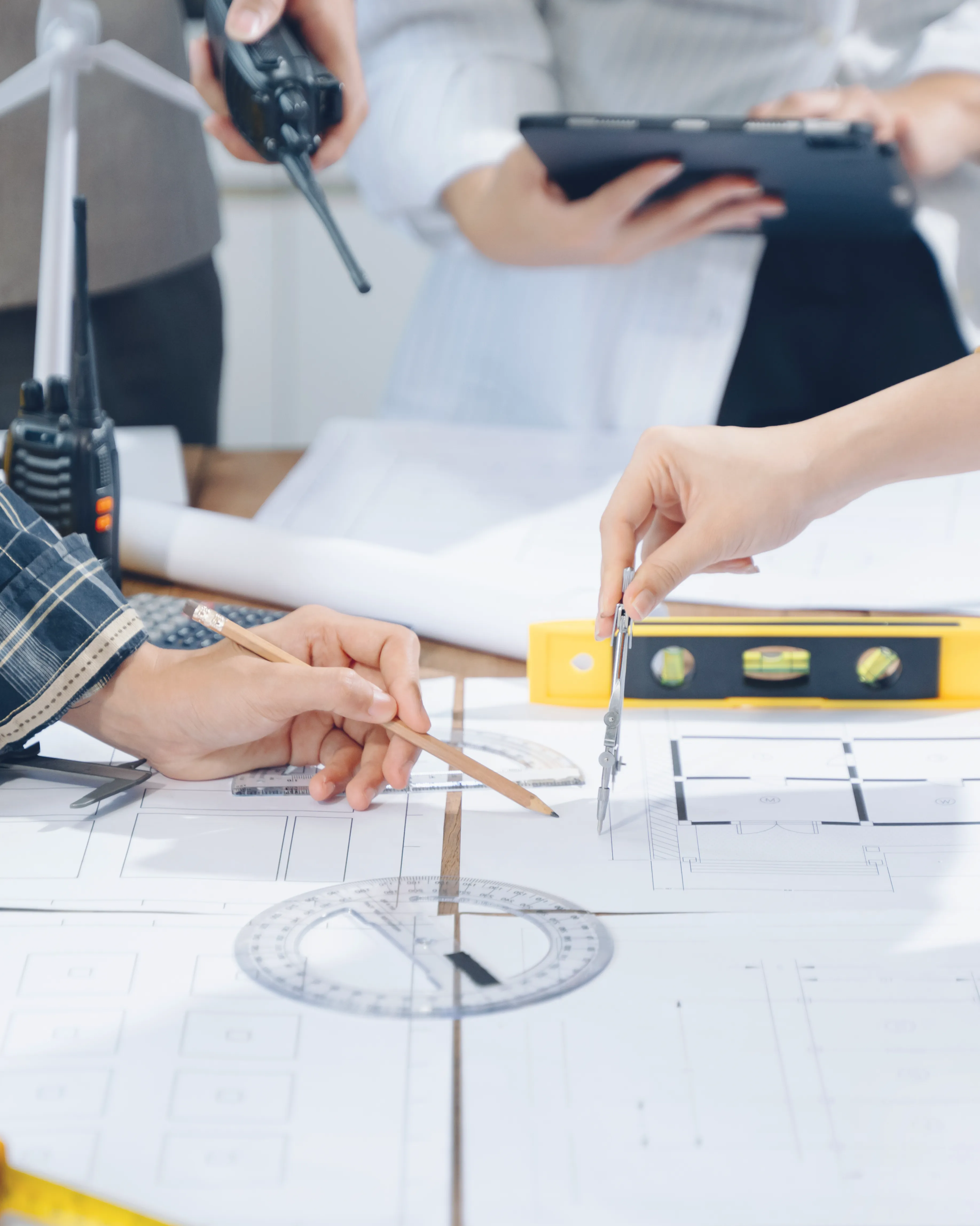 Two people working on architectural blueprints with drawing tools including a pencil, compass, and protractor while another person holds a walkie-talkie and a tablet in the background.