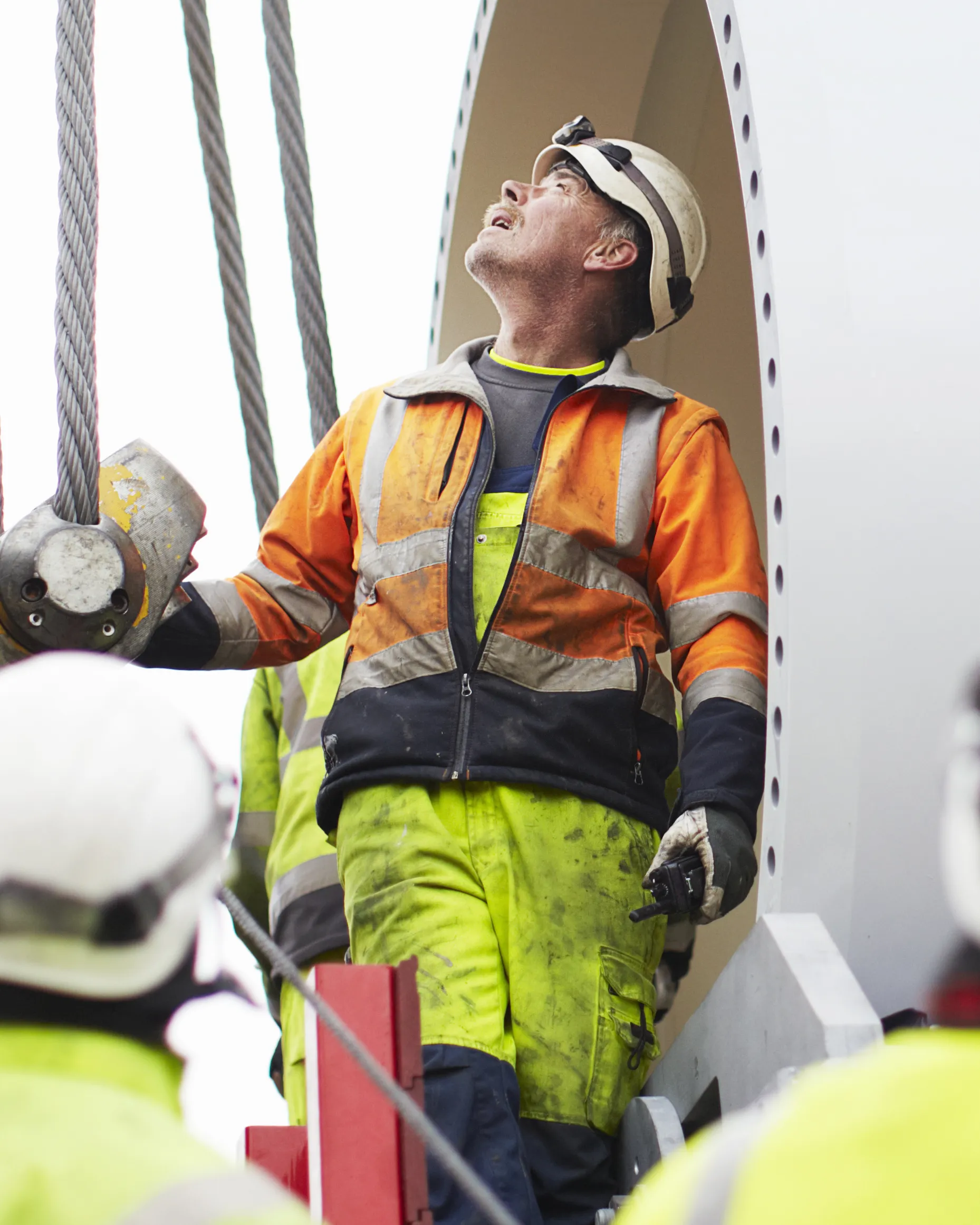 Engineer in a hard hat and high-visibility clothing looking up while working with cables on industrial equipment.