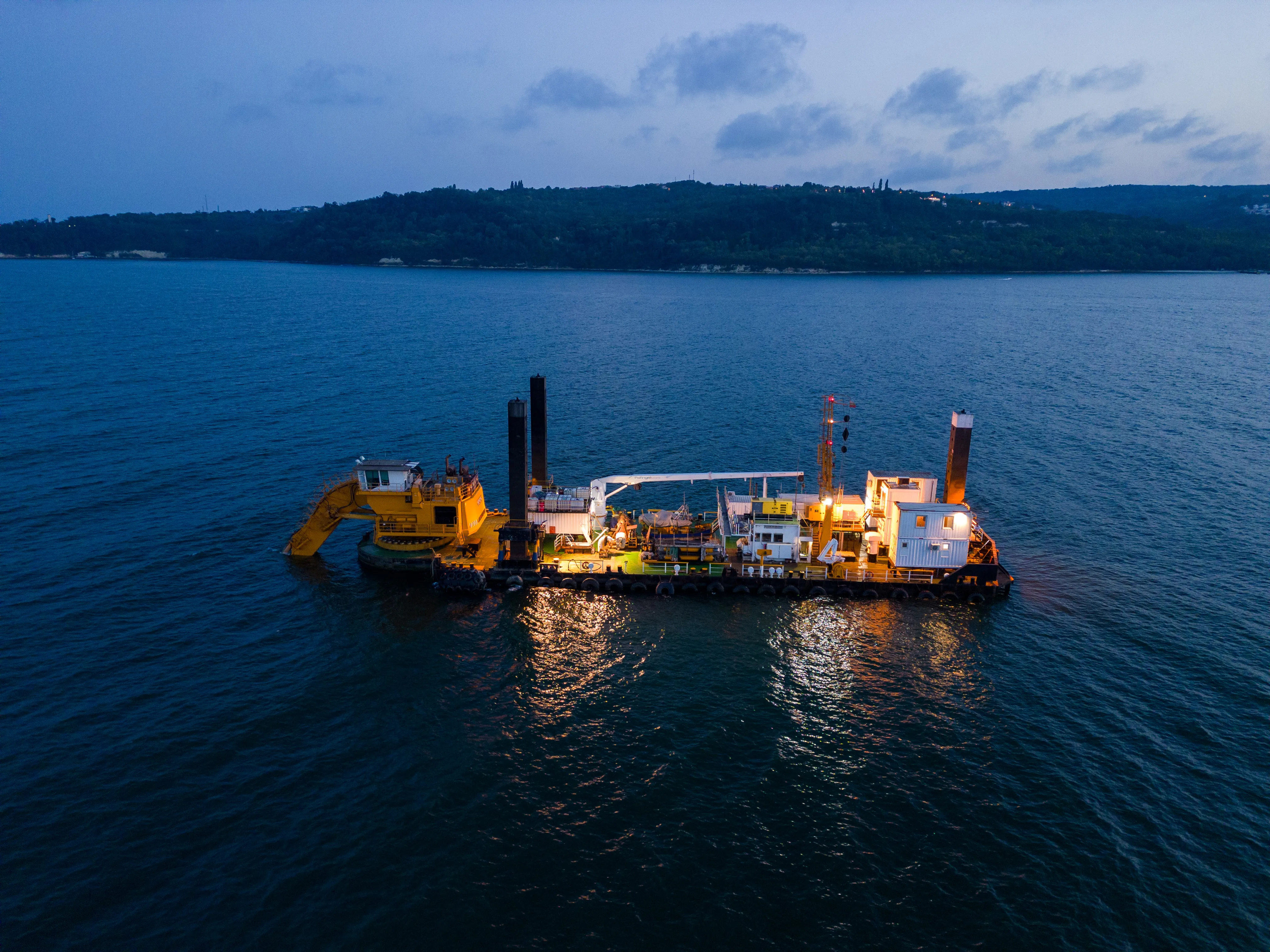 Aerial view of an illuminated yellow dredge vessel floating on calm water near a forested coastline at dusk.